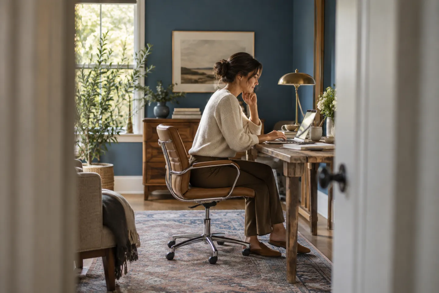 A consultant scheduling a client meeting from a quiet blue-walled home office, side view through a doorway
